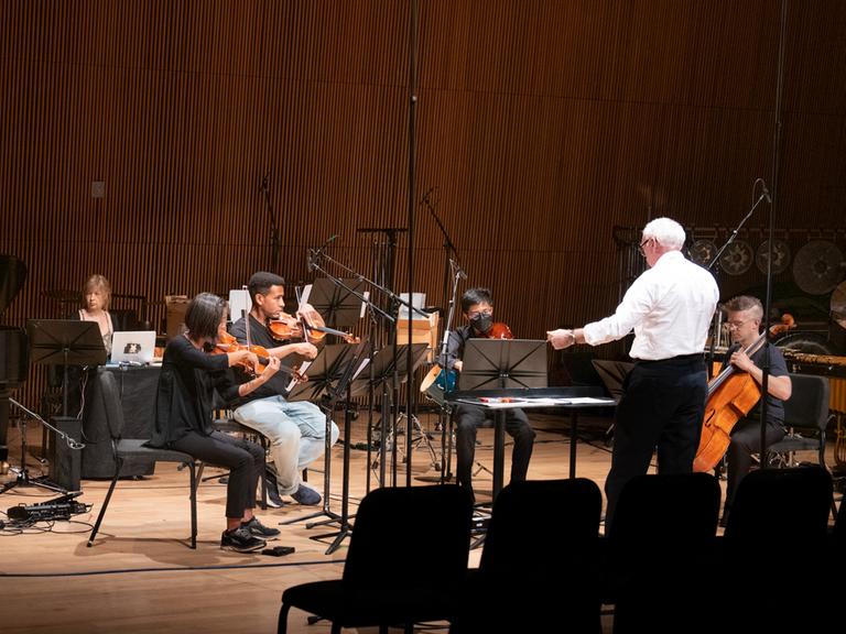 Ein Mann in weißem Hemd dirigiert ein Ensemble von 5 Musikern auf einer Bühne mit braunem Parkettboden