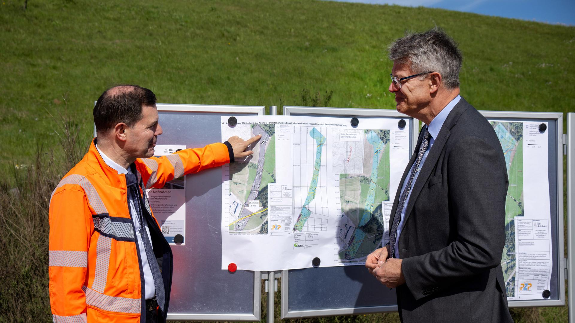 Bundesverkehrsminister Schnieder steht mit einem Vertreter der Autobahn GmbH, der eine orangefarbene Jacke trägt, vor einer Informationstafel mit Karten zum aktuellen Stand der Baumaßnahmen an A1 in der Eifel.