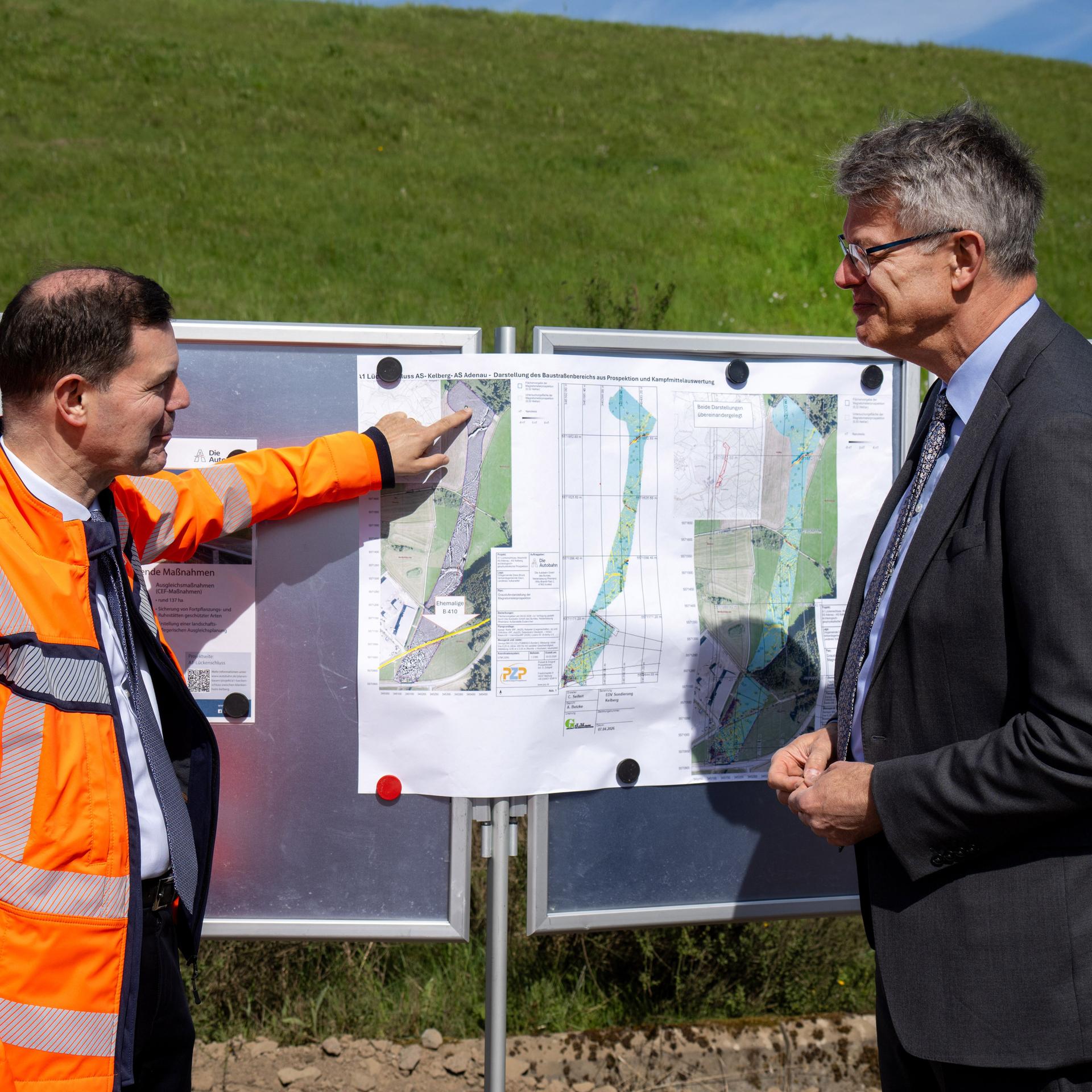 Bundesverkehrsminister Schnieder steht mit einem Vertreter der Autobahn GmbH, der eine orangefarbene Jacke trägt, vor einer Informationstafel mit Karten zum aktuellen Stand der Baumaßnahmen an A1 in der Eifel.
