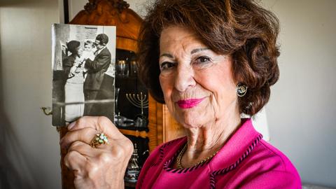 Irene Bindel trägt eine pinke Bluse und einen pinken Lippenstift. Sie hält eine Schwarzweißfotografie in die Kamera, die sie als Kind mit ihren Eltern zeigt.