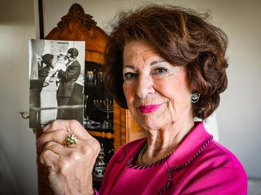 Irene Bindel trägt eine pinke Bluse und einen pinken Lippenstift. Sie hält eine Schwarzweißfotografie in die Kamera, die sie als Kind mit ihren Eltern zeigt. Irene Bindel trägt eine pinke Bluse und einen pinken Lippenstift. Sie hält eine Schwarzweißfotografie in die Kamera, die sie als Kind mit ihren Eltern zeigt.