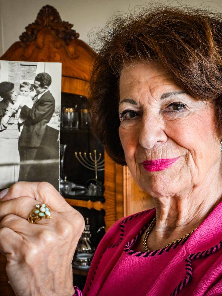 Irene Bindel trägt eine pinke Bluse und einen pinken Lippenstift. Sie hält eine Schwarzweißfotografie in die Kamera, die sie als Kind mit ihren Eltern zeigt. Irene Bindel trägt eine pinke Bluse und einen pinken Lippenstift. Sie hält eine Schwarzweißfotografie in die Kamera, die sie als Kind mit ihren Eltern zeigt.