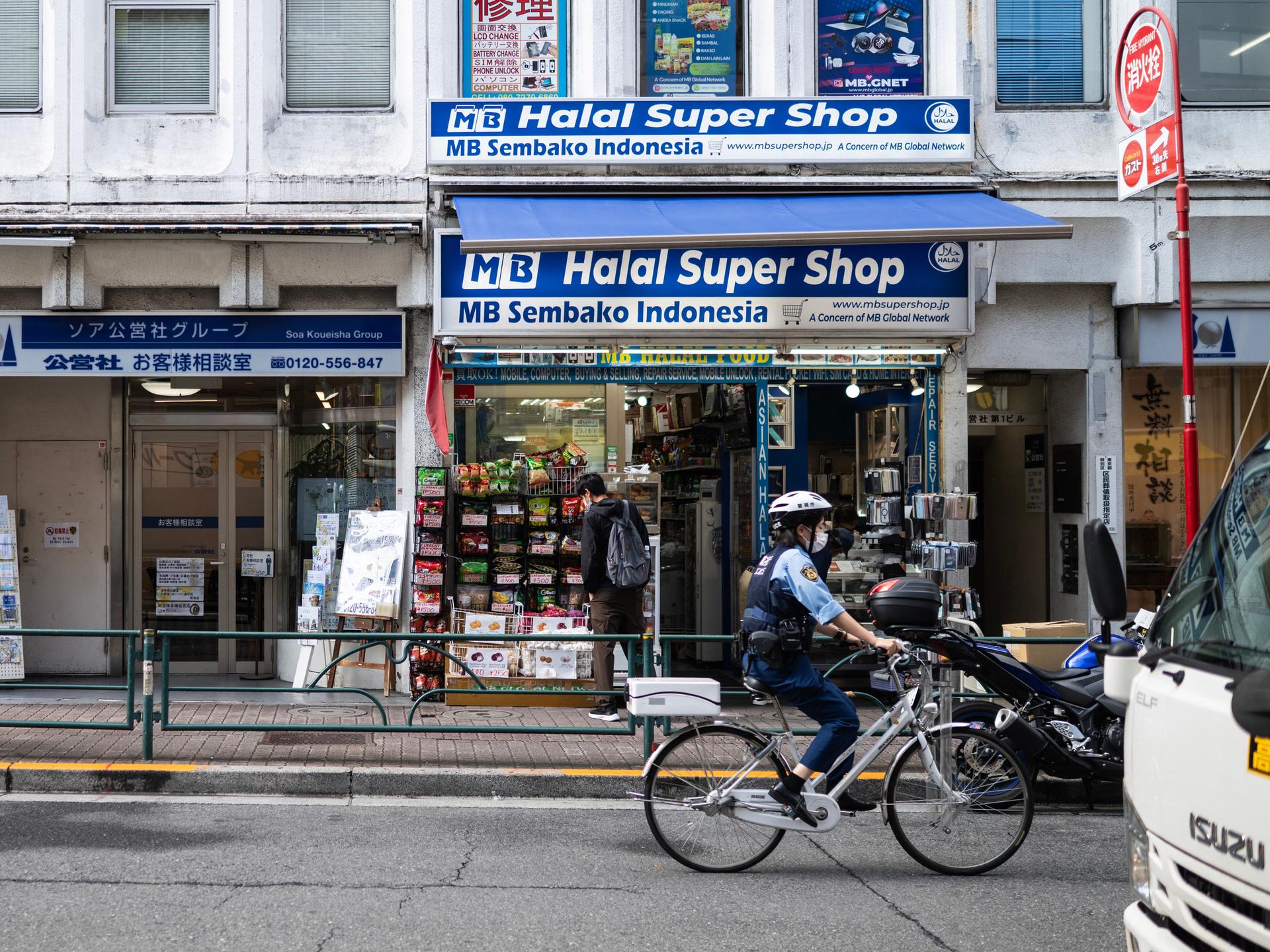Ein Polizist fährt mit dem Fahrrad an einem Geschäft im Tokioter Stadtviertel Okubo vorbei. Das Banner am Geschäft verät, dass die Inhaber aus Indonesien stammen und Halal Produkte verkaufen.