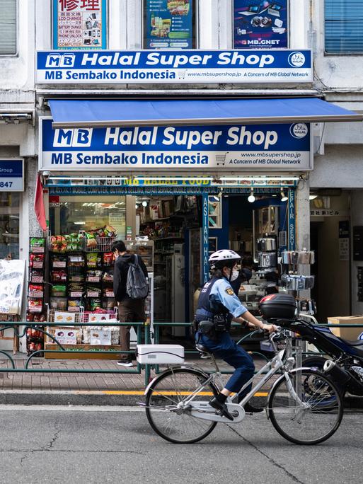 Ein Polizist fährt mit dem Fahrrad an einem Geschäft im Tokioter Stadtviertel Okubo vorbei. Das Banner am Geschäft verät, dass die Inhaber aus Indonesien stammen und Halal Produkte verkaufen.