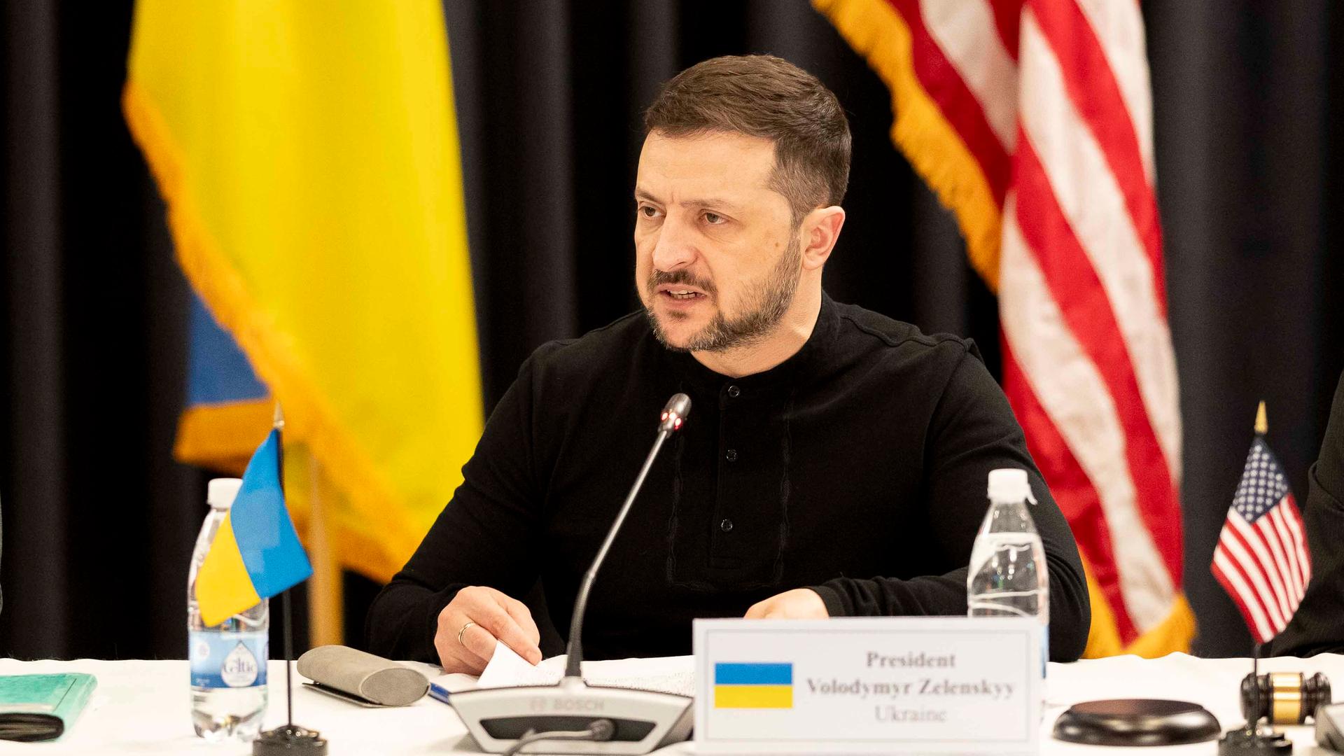 Der ukrainische Präsident Selenskyj sitzt beim Treffen der Ukraine-Partner in Ramstein an einem Tisch, neben sich die Flagge der Ukraine sowie die US-Flagge, und hält mit wütendem Gesichtsausdruck eine Rede.