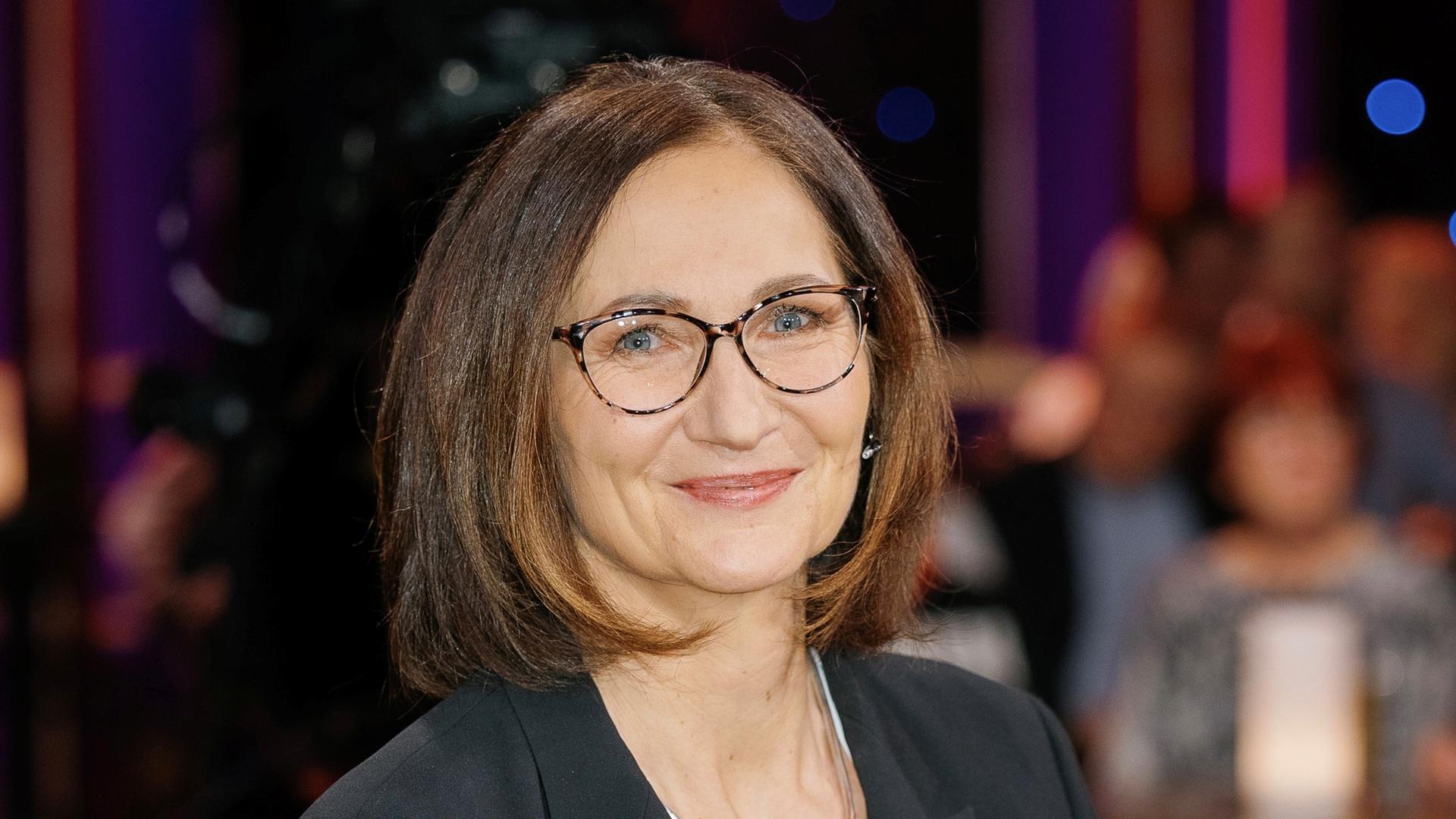 Eine Frau mit brünettem Haar und Brille blickt lächelnd in die Kamera. Sie trägt ein schwarzes Jacket über einem weißen Shirt und eine silberne Kette. Im unscharfen Hintergrund sind Menschen zu sehen, die an Tischen sitzen.