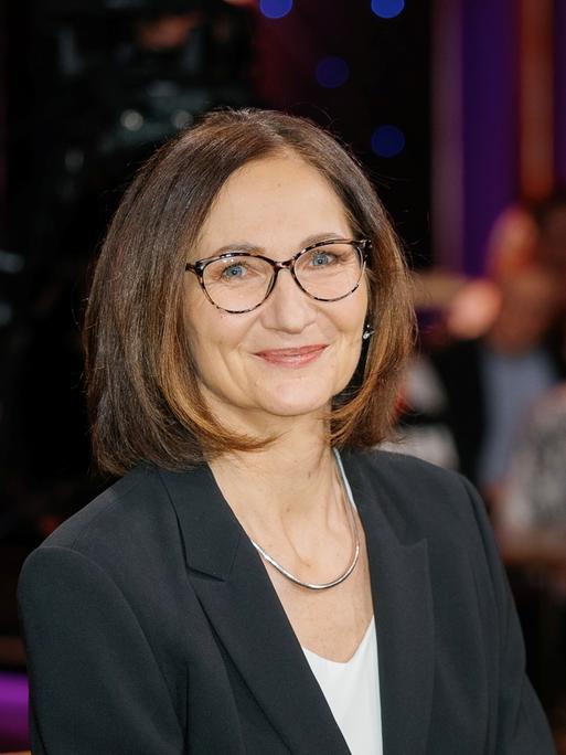 Eine Frau mit brünettem Haar und Brille blickt lächelnd in die Kamera. Sie trägt ein schwarzes Jacket über einem weißen Shirt und eine silberne Kette. Im unscharfen Hintergrund sind Menschen zu sehen, die an Tischen sitzen.