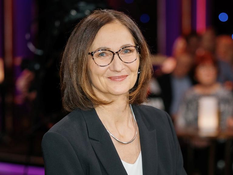 Eine Frau mit brünettem Haar und Brille blickt lächelnd in die Kamera. Sie trägt ein schwarzes Jacket über einem weißen Shirt und eine silberne Kette. Im unscharfen Hintergrund sind Menschen zu sehen, die an Tischen sitzen.