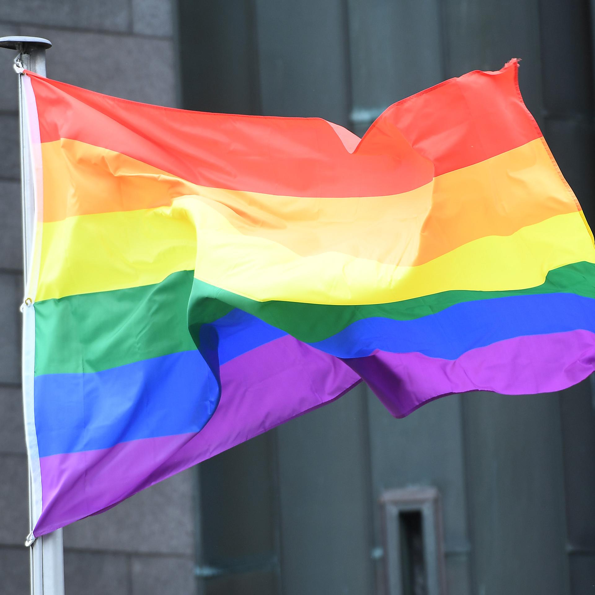 Eine Regenbogenfahne weht vor einem Gebäude im Wind. 