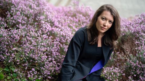 Porträt von Maria Reva vor einer blühenden Hecke. Sie hat dunkles schulterlanges Haar und trägt einen blauen Blazer.