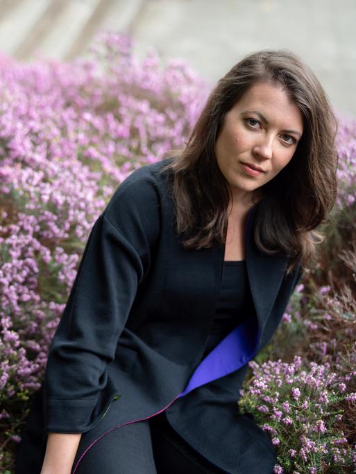 Porträt von Maria Reva vor einer blühenden Hecke. Sie hat dunkles schulterlanges Haar und trägt einen blauen Blazer. Porträt von Maria Reva vor einer blühenden Hecke. Sie hat dunkles schulterlanges Haar und trägt einen blauen Blazer.