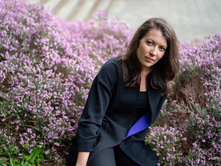 Porträt von Maria Reva vor einer blühenden Hecke. Sie hat dunkles schulterlanges Haar und trägt einen blauen Blazer. Porträt von Maria Reva vor einer blühenden Hecke. Sie hat dunkles schulterlanges Haar und trägt einen blauen Blazer.