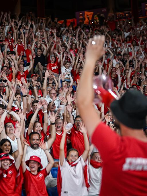 Schweizer Fans beim Eröffnungsspiel gegen Norwegen. Die Zuschauerinnen und Zuschauer heben fröhlich die Hände zu La Ola, sie tragen Schweizer Trikots. 