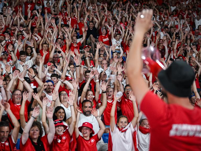 Schweizer Fans beim Eröffnungsspiel gegen Norwegen. Die Zuschauerinnen und Zuschauer heben fröhlich die Hände zu La Ola, sie tragen Schweizer Trikots. 