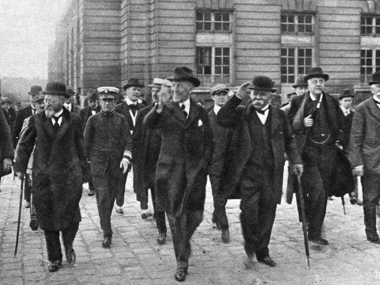 Schwarzweiß Kulisse vor der mehrere Männer in schwarzen Anzügen und Hüten laufen.
