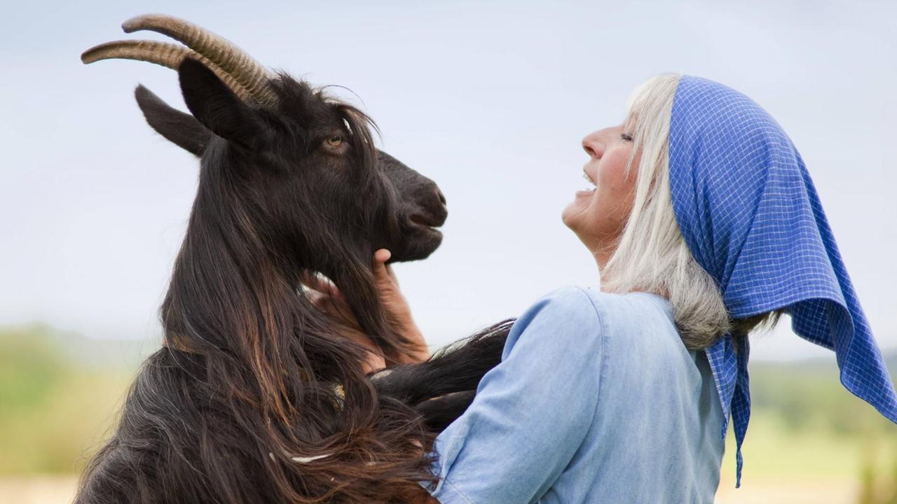 Ein Ziegenbock hat seine Vorderhufe auf die Schultern einer Frau gelegt, sie lacht ihn an.
