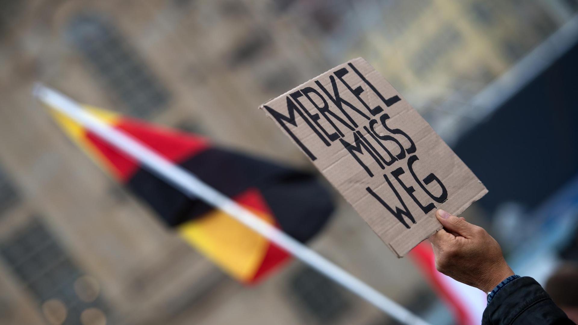 Eine Hand hält ein Schild mit der Aufschrift "Merkel muss weg" in die Höhe, dahinter eine deutsche Flagge.