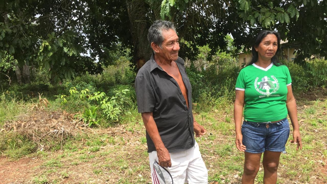Gemeindeleiterin Amelia Praga Cabral  im knallgrünen T-Shirt und kurzen Jeans steht mit einem Mann vor einem Wald auf einer Wiese.