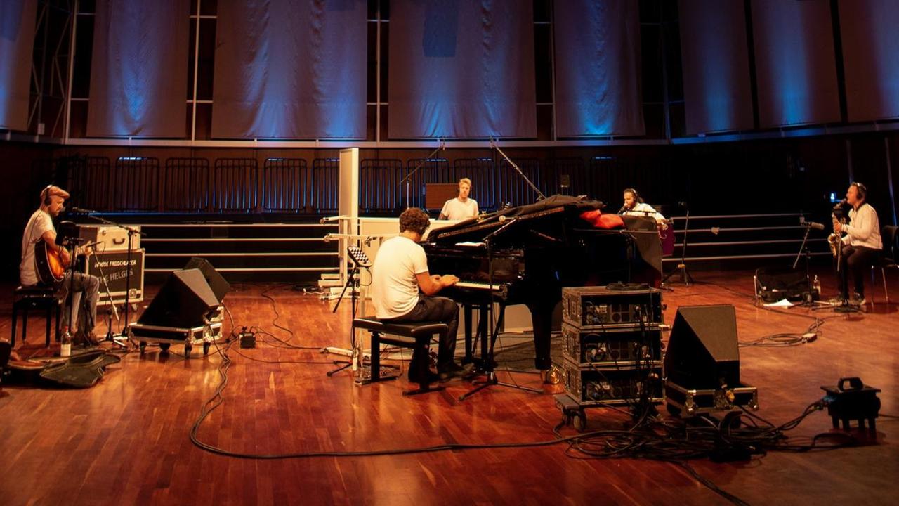 Fünf junge Männer sitzen in einem bläulich ausgeleuchteten Konzertsaal und spielen Instrumente.
