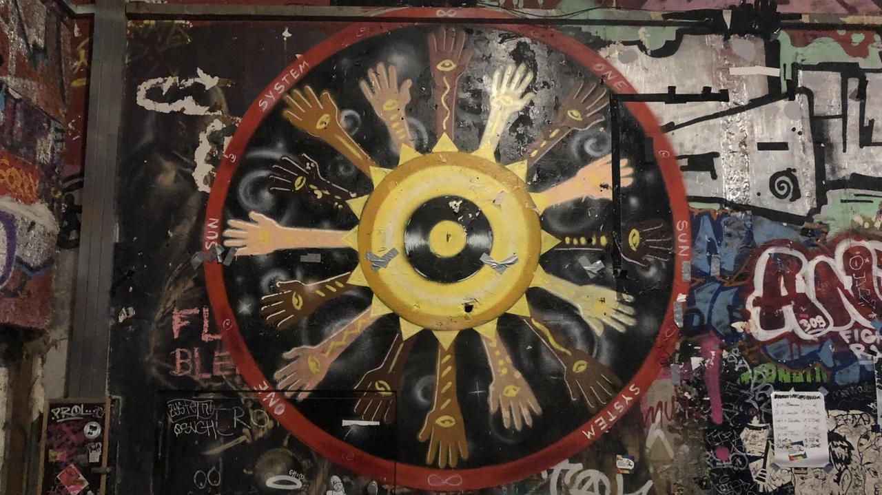 Das Bild zeigt ein Graffito, dessen Mitte ein Vinyl-Langspielplatte bildet. Kreisförmig angeordnet  sind Hände, die verschiedene Ethnien symbolisieren.