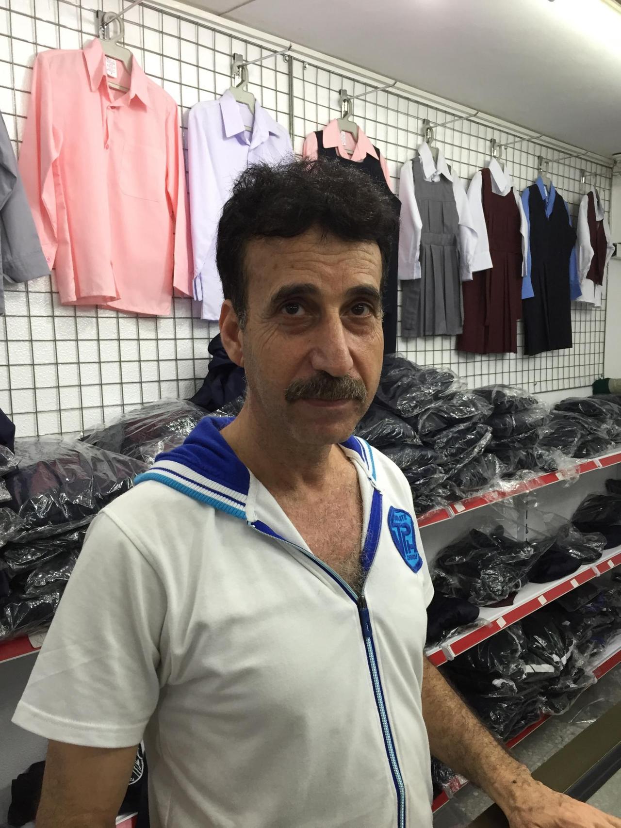 Samir Sada, Betreiber eines Schulfachgeschäfts, in seinem Laden, vor Regalen und Bügeln mit Kleidung.