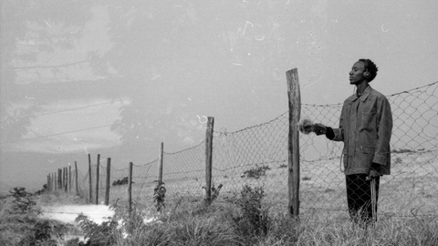 Schwarz-Weiss Fotografie von Joseph Kamaru, der mit Kopfhörern an einem Maschendrahtzaun steht.