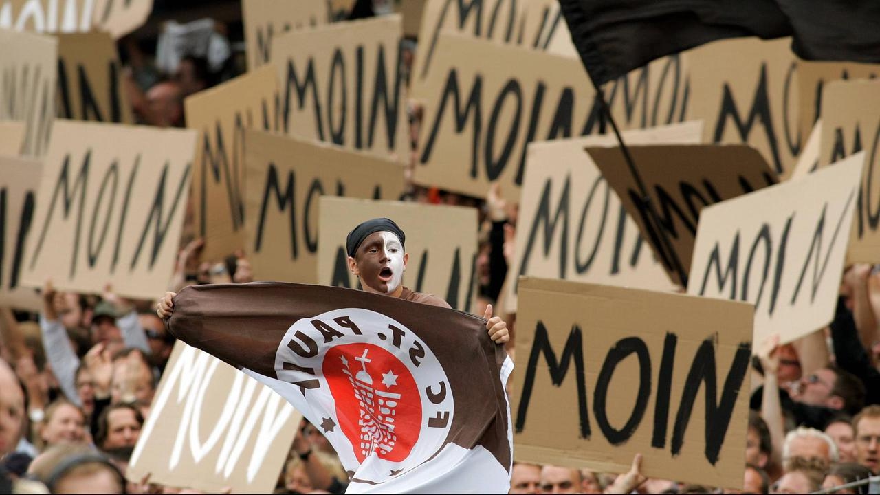 St. Pauli Fan mit Gesichtsbemalung und Vereinsfahne umringt von Fanplakaten auf denen "Moin" steht.