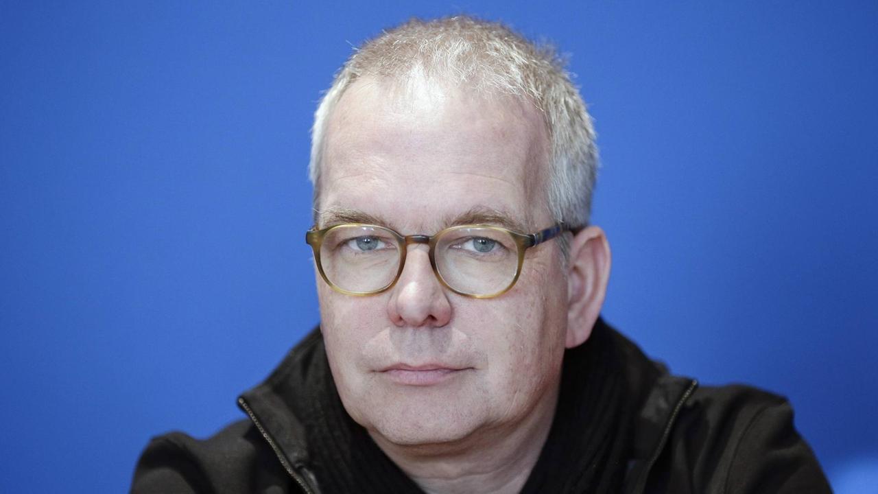 Ein Mann mit etwas schütterem Haar und Brille blickt vor einem blauen Hintergrund in die Kamera.
