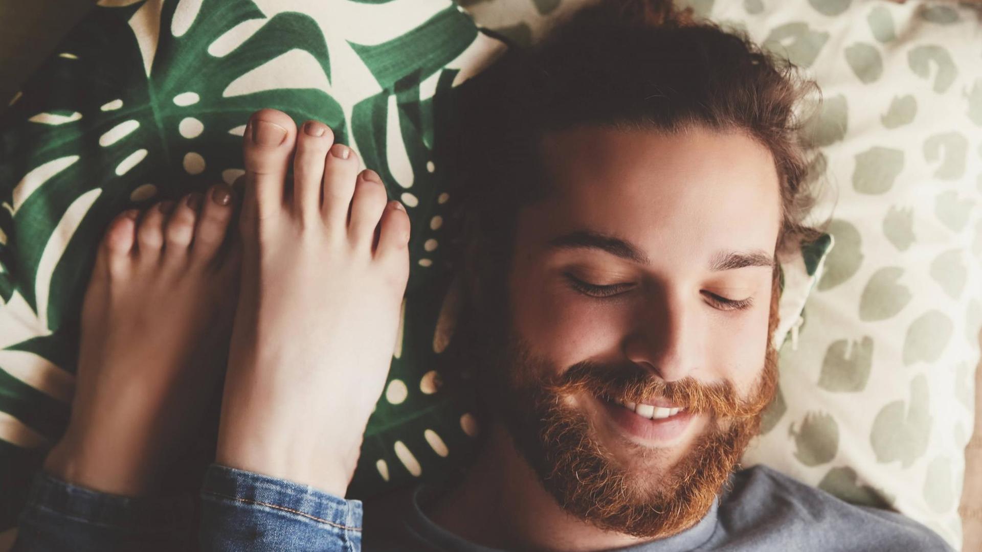 Ein lächelnder Mann liegt auf dem Bett, neben ihm Füße einer Frau.