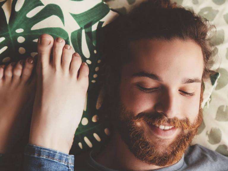 Ein lächelnder Mann liegt auf dem Bett, neben ihm Füße einer Frau.