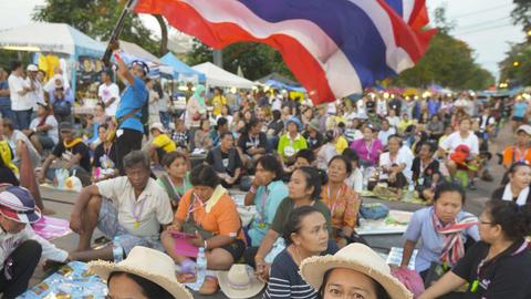 Eine Menschenmenge hockt auf dem Boden, vorne zwei Demonstranten hervorgehoben, im Hintergrund wird eine thailändische Flagge geschwenkt.