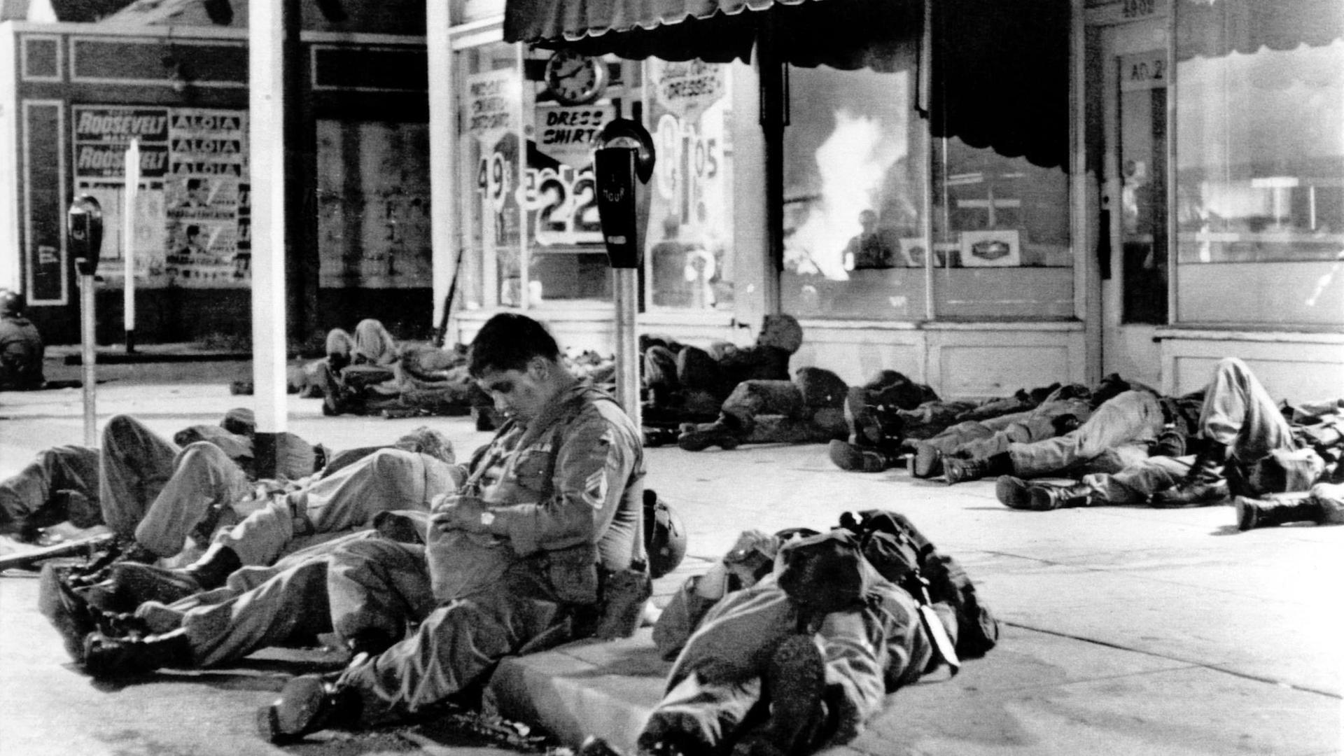 Erschöpfte Soldaten der Nationalgarde schlafen auf den Bürgersteigen im Watts-Distrikt nach Rassenunruhen in Los Angelers. In den Schaufenstern der Geschäfte spiegeln sich die Flammen eines brennenden Gebäudes.