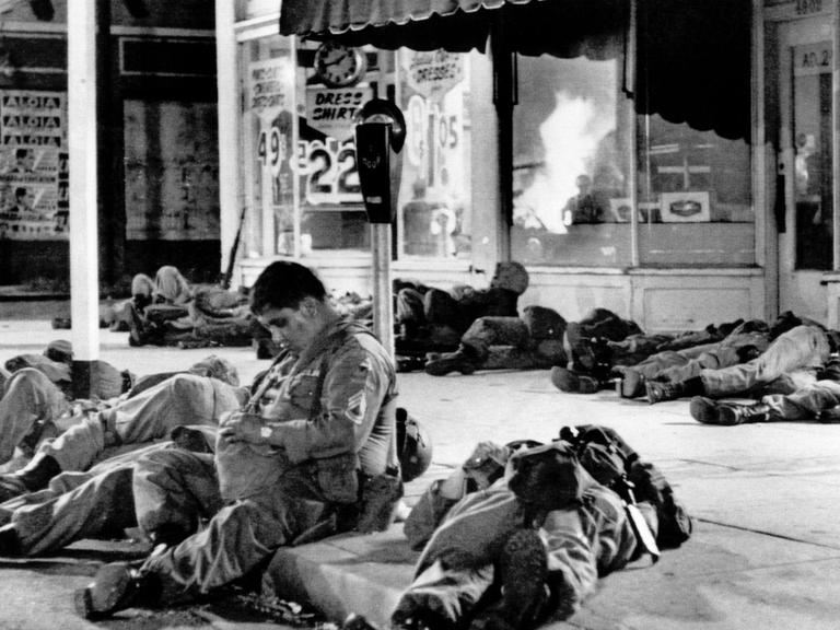 Erschöpfte Soldaten der Nationalgarde schlafen auf den Bürgersteigen im Watts-Distrikt nach Rassenunruhen in Los Angelers. In den Schaufenstern der Geschäfte spiegeln sich die Flammen eines brennenden Gebäudes.