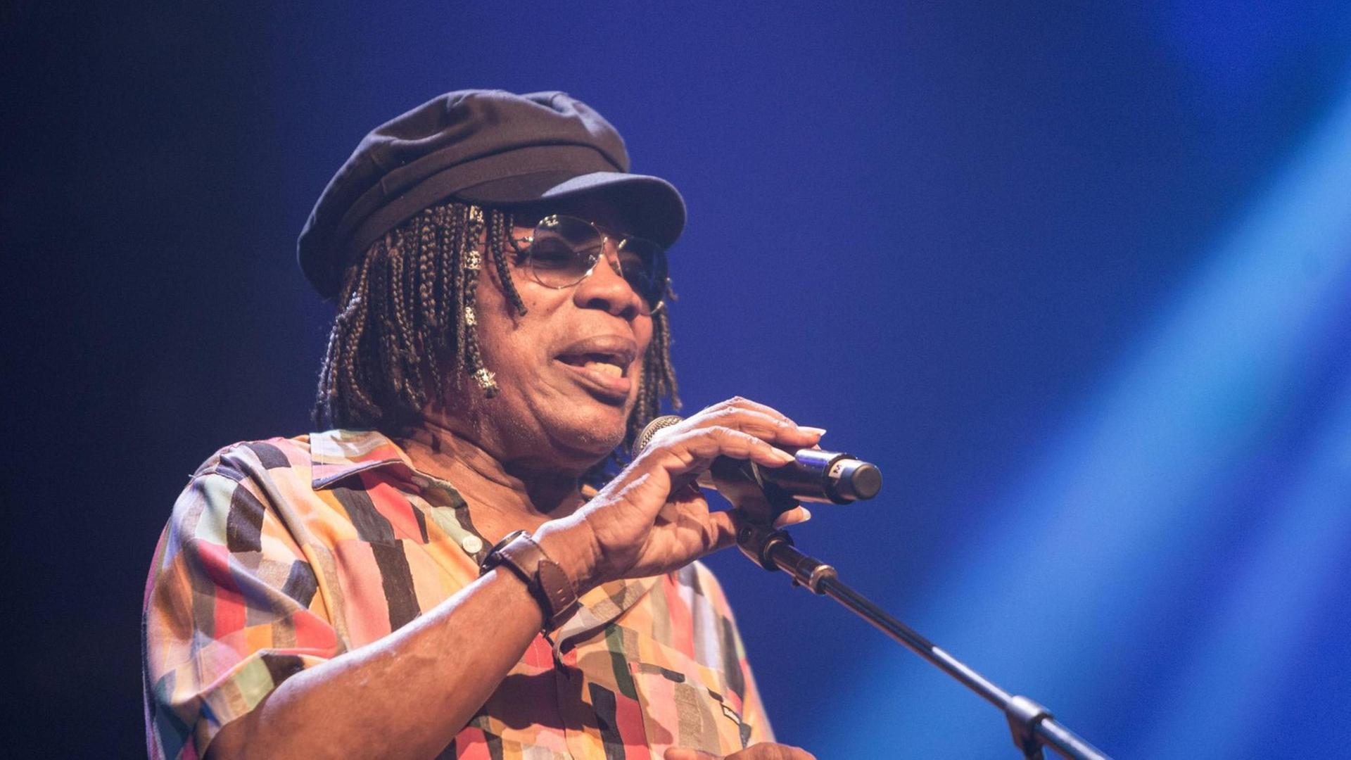 Milton Nascimento trägt eine Brille und eine Mütze und singt auf einer Bühne in ein Mikrofon.