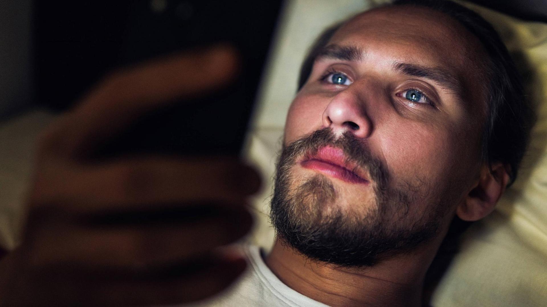 Ein Mann liegt im Bett und schaut auf sein Smartphone-Display.