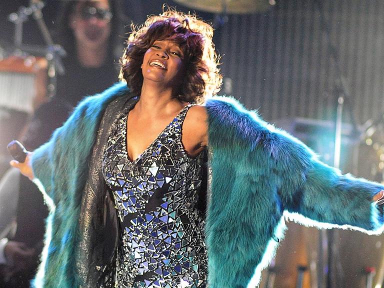 Whitney Houston auf der Bühne, lacht und streckt die Hände aus. Sie trägt ein silbernes Glitzerkleid und darüber einen grünen Pelzmantel.