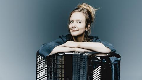 Eine mittelalte Frau stützt sich mit verschränkten Armen auf ein Akkordeon und lächelt in die Kamera.