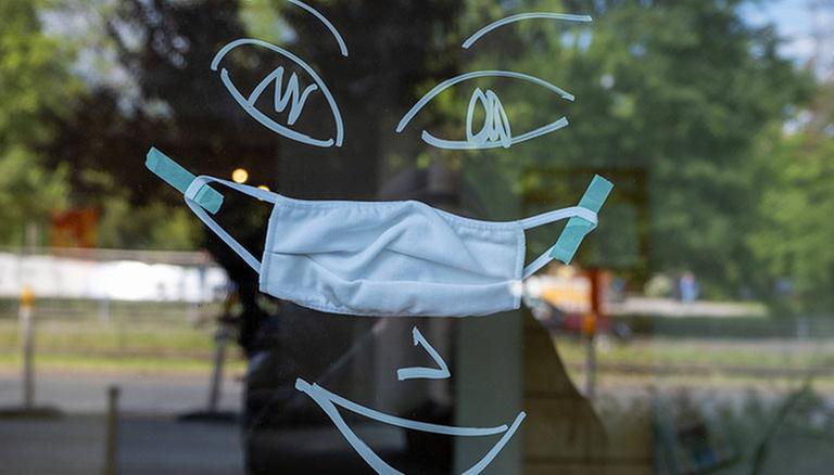 An ein Schaufenster gemaltes freundliches Gesicht mit einem Mund-Nasen-Schutz. In Berlin gilt seit kurzen eine Pflicht zum Tragen von Gesichtsbedeckungen im Einzelhandel. Coronakrise - Freundliches Gesicht mit Mundschutz *** Friendly face painted on a shop window with a mouth Noses Protection In Berlin, there has recently been a requirement to wear face coverings in retail stores Coronakrise Friendly face with mouthguard