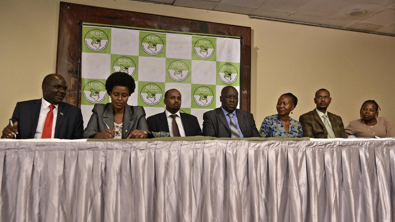 Das Bild zeigt die siebenköpfige Wahlkommission von Kenia, nebeneinander an einem Tisch sitzend. An dritter Stelle von rechts sitzt Roselyn Akombe, die aus der Kommission zurückgetreten und in die USA geflohen ist.