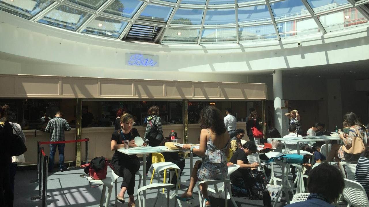 Farbfoto einer Bar in Cannes, Südfrankreich, in der man während der Filmfestspiele etwas essen kann. Mehrere Besucher sitzen an Tischen oder bestellen an einer Theke etwas