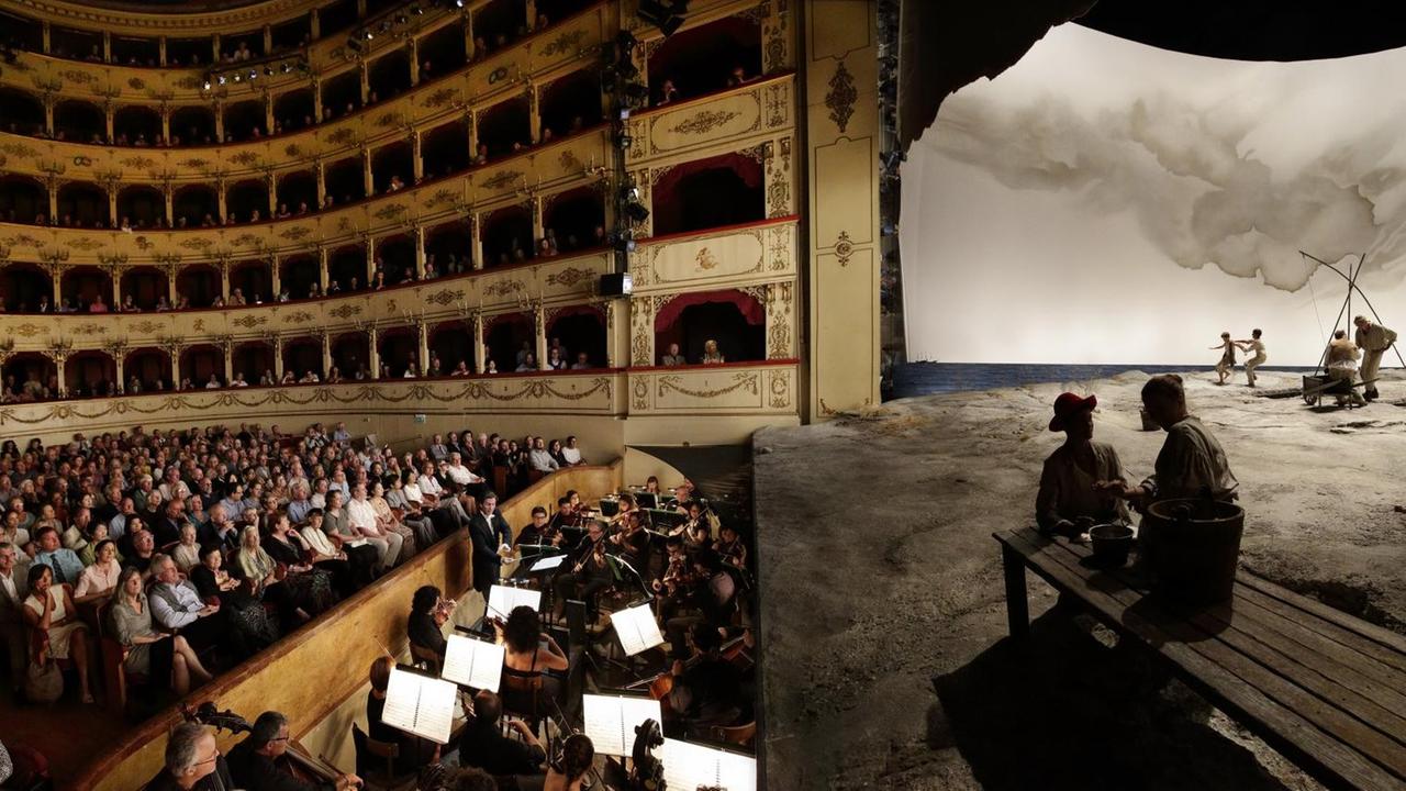 Blick von der Bühnenkante zu beiden Seiten des Raumes. Links sieht man das Publikum, das in einem historischen, italienischen Opernsaal mit vielen Logen Richtung Bühne schaut, die auf der rechten Seite zu sehen ist und eine karge Landschaft darstellt.