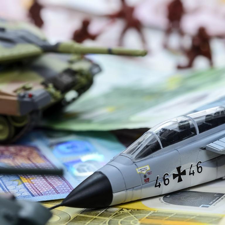 Das Symbolbild zeigt das Modell eines Panzers und eines Militärjets sowie Geldscheine.