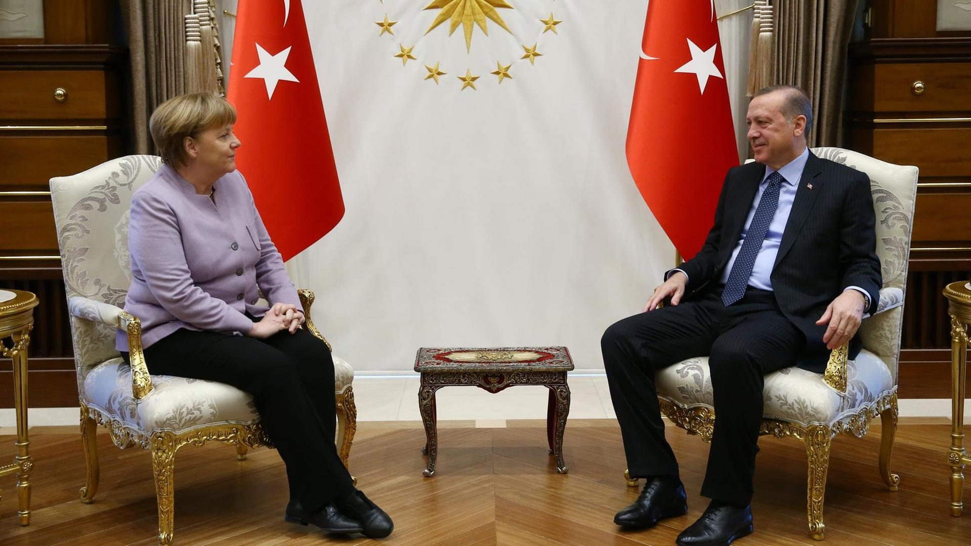 Die beiden sitzen sich in Stühlen gegenüber und lächeln, dahinter stehen zwei türkische Fahnen.
