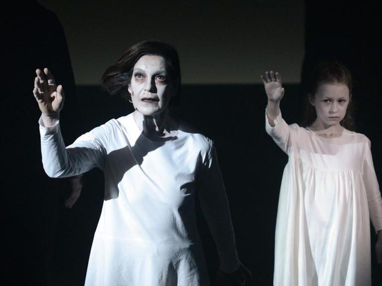Eine Frau und ein Mädchen in weißen langen Kleidern stehen angestrahlt auf sonst dunkler Bühne und heben die Arme.