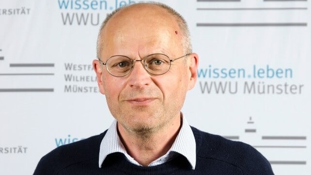 Ein Mann mit Brille, in Hemd und Pullover, steht vor einem Plakat der Universität Münster und schaut leicht stirnrunzelnd, aber lächelnd in die Kamera.