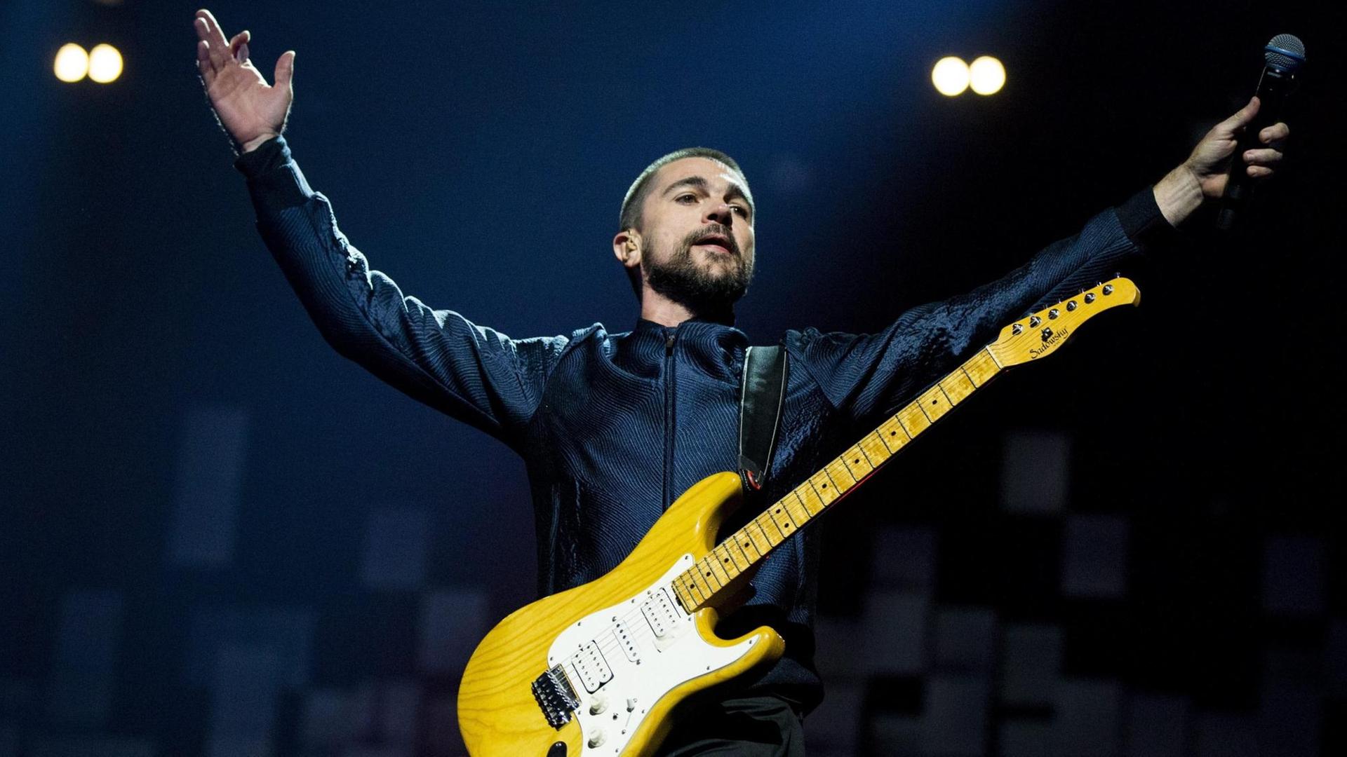 Juanes auf der Bühne, reißt die Arme hoch und trägt eine gelbe E-Gitarre