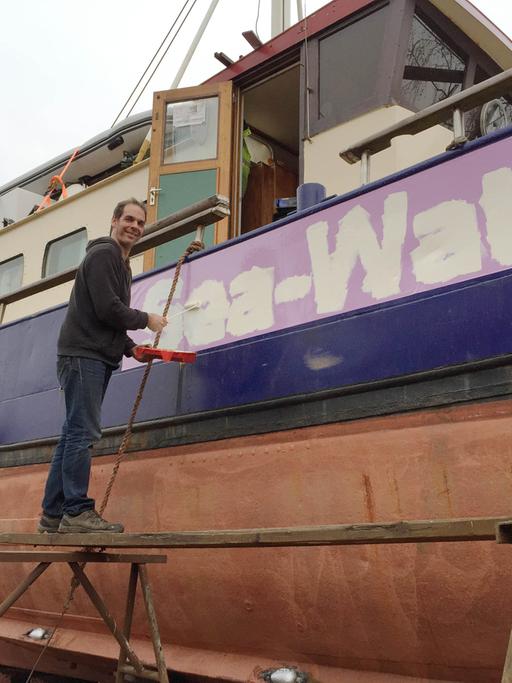 Harald Höppner steht auf einem Holzbrett vor einem Schiff, mit Malerutensilien in der Hand, auf dem Schiff steht "Sea-Watch" geschrieben.