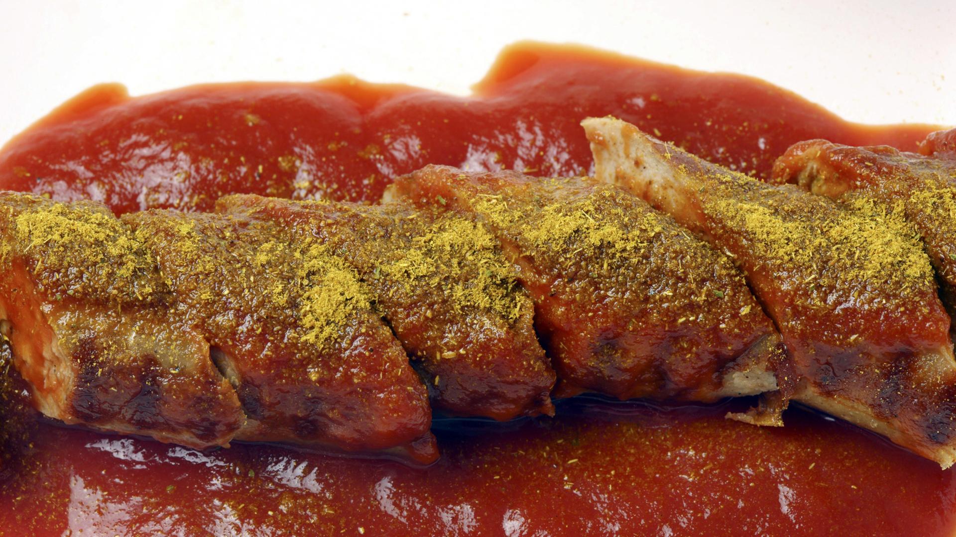 Currywurst mit roter Soße, Nahaufnahme: Auf der Wurst ist gelbes Currypulver zu sehen.