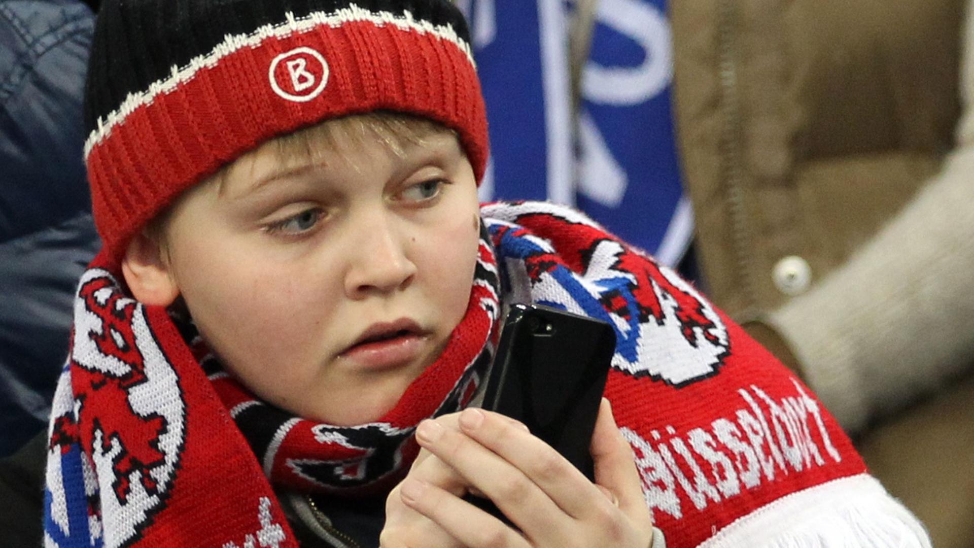 Ein junger Düsseldorfer Fan hält gespannt ein Mobiltelefon in der Hand.