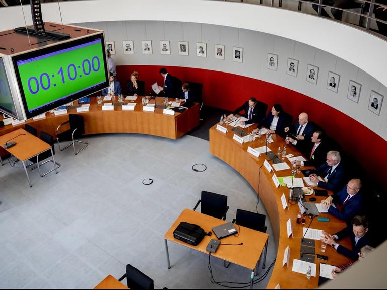 Das Bild ist vom Besucherrang aus gemacht; man sieht unten die Teilnehmer an halbrunden Tischen und oben Zuschauer auf der Tribüne. Von der Decke hängt eine viereckige Digitaluhr herab.